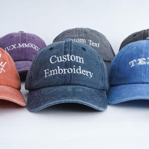 Op de afbeelding: Een collectie van vijf baseballpetten in diverse kleuren, waaronder oranje, paars, donkerblauw, grijs en lichtblauw. Elke pet is voorzien van wit geborduurde tekst, waaronder de woorden Firefly, IV.X.MMXXV, Custom Embroidery en TEXT.