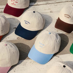 Peut inclure: Une collection de casquettes de baseball avec des calottes couleur crème et des visières de couleurs contrastées. Chaque casquette présente du texte brodé incluant Margarita Est 2004, Embroidery Here, Grandpa Est 2025, Custom text et Logo.
