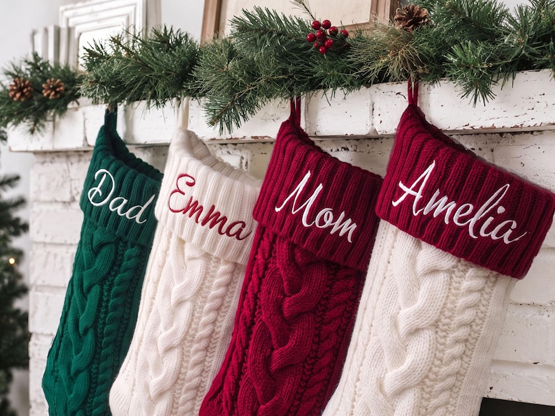 May include: Four knit Christmas stockings featuring a cable knit pattern. The first is dark green with white embroidery reading Dad, the second is white with red embroidery reading Emma, the third is red with white embroidery reading Mom, and the fourth is white with red embroidery reading Amelia.