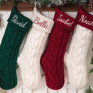 May include: Four cable-knit Christmas stockings hanging in a row. The first is dark green with the name David, the second is cream with the name Bella, the third is deep red with the name Isabel, and the fourth is cream with the name Noah.
