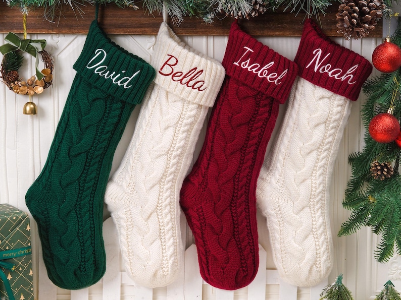 May include: Four cable-knit Christmas stockings hanging in a row. The first is dark green with the name David, the second is cream with the name Bella, the third is deep red with the name Isabel, and the fourth is cream with the name Noah.
