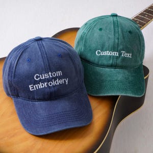 May include: A blue baseball cap with the white embroidered text Custom Embroidery and a green baseball cap with the white embroidered text Custom Text. Both caps feature a washed fabric finish, a curved brim, and a six-panel construction.