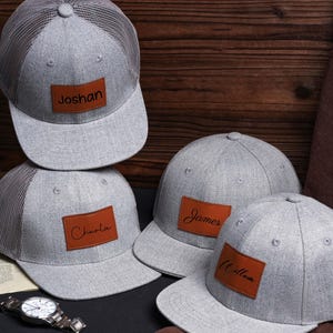 May include: Four gray baseball caps with brown leather patches. Each patch has a name in black script: Joshan, Charlie, James, and William. The caps have a curved brim and a button on top. One cap has a mesh back.
