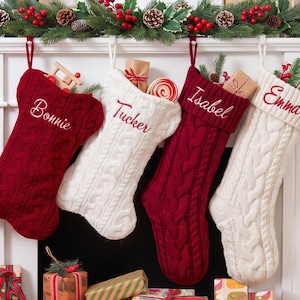 Könnte beinhalten: Vier Weihnachtsstrümpfe aus Zopfstrick hängen in einer Reihe und wechseln sich in den Farben Dunkelrot und Cremeweiß ab. Jeder Strumpf ist mit einer personalisierten Stickerei in Schreibschrift versehen, die die Namen Bonnie, Tucker, Isabel und Emma zeigt.