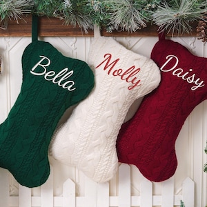 May include: Three bone-shaped Christmas stockings made of cable-knit fabric in dark green, cream, and deep red. Each stocking features personalized embroidery: the green one says Bella, the cream one says Molly, and the red one says Daisy.