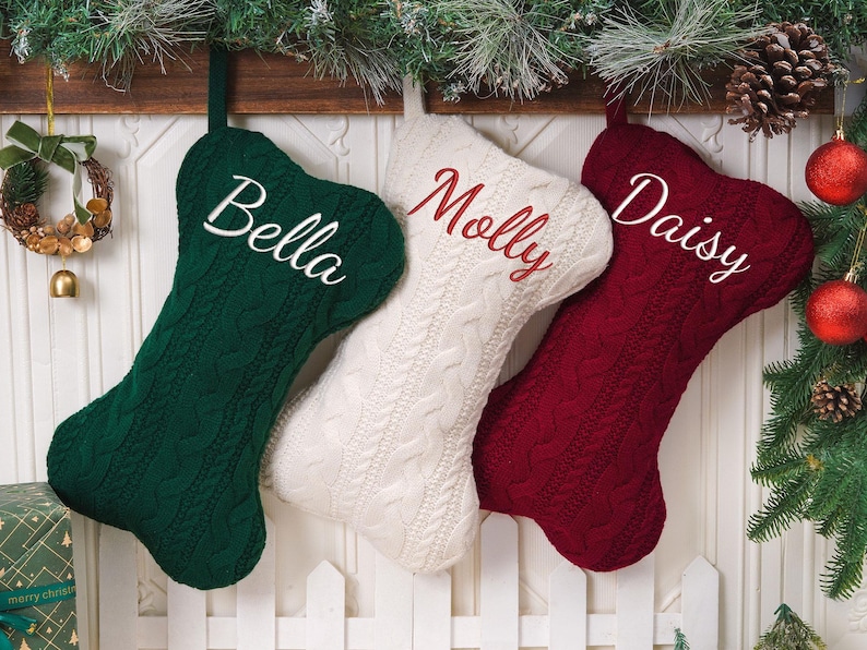 May include: Three bone-shaped Christmas stockings made of cable-knit fabric in dark green, cream, and deep red. Each stocking features personalized embroidery: the green one says Bella, the cream one says Molly, and the red one says Daisy.