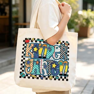 May include: A cream-colored canvas tote bag featuring a large, colorful graphic print that spells out the word KINDERGARTEN. The letters are decorated with various patterns, including checkers, stripes, and stars, surrounded by small school-themed icons.