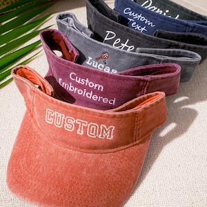 May include: A stack of five adjustable sun visors in various colors including orange, maroon, gray, navy blue, and black. Each visor features white embroidered text such as CUSTOM, Custom Embroidered, Lucas with a tennis racket, Pete, and Daniel.