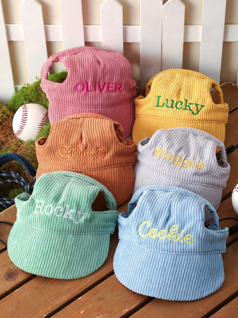 May include: Six corduroy pet sun visors in pink, yellow, orange, grey, mint green, and light blue. Each visor features an embroidered name: Oliver, Lucky, Coco, Maggie, Rocky, and Cookie. The hats have ear holes and a structured brim design.