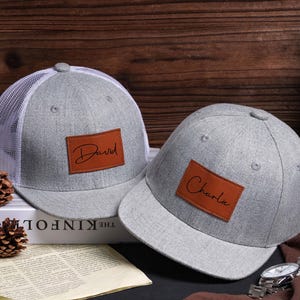 May include: Two gray baseball caps with brown leather patches. One cap has a white mesh back and the name "David" on the patch. The other cap has the name "Charlie" on its patch. Both caps are on a wooden surface.