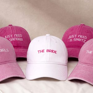 Puede incluir: Un conjunto de gorras de béisbol rosas y blancas. Las gorras presentan bordados con textos como "THE GIRLS", "JUST NEED SANGRIA", "JUST NEED A SPRITZ" y "THE BRIDE".