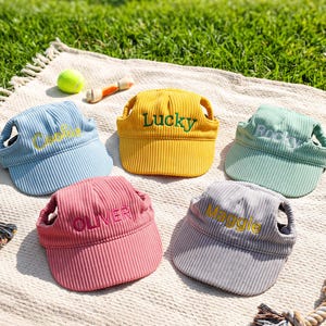 May include: Five corduroy pet visors in light blue, yellow, mint green, pink, and gray. Each visor features embroidered names: Cookie in yellow, Lucky in green, Rocky in white, OLIVER in pink, and Maggie in yellow, with ear holes on the sides.