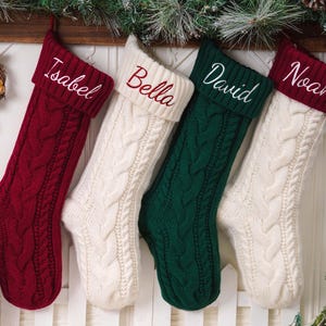 May include: Four cable-knit Christmas stockings hanging in a row. The first is deep red with the name Isabel, the second is cream with Bella, the third is dark green with David, and the fourth is cream with a deep red cuff featuring the name Noah.