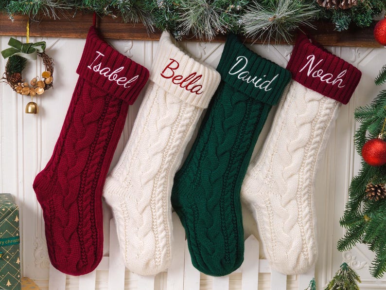May include: Four cable-knit Christmas stockings hanging in a row. The first is deep red with the name Isabel, the second is cream with Bella, the third is dark green with David, and the fourth is cream with a deep red cuff featuring the name Noah.