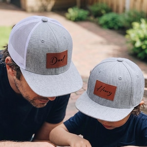 May include: Two grey baseball caps with brown leather patches. One cap has "Dad" in script, the other has "Henry". The caps have a grey brim and a white mesh back. The caps are worn by a person and a child.