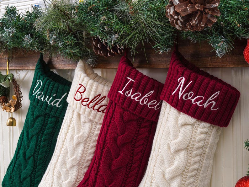 May include: Four cable-knit Christmas stockings hanging in a row. The first is dark green with the name David, the second is cream with Bella, the third is deep red with Isabel, and the fourth is cream with a deep red cuff featuring the name Noah.