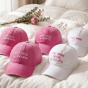 May include: Five baseball caps in pink and white with embroidered text. The pink caps feature the phrases Just Need A Margarita, Just Need A Spritz, and Just Need A Sangria. The white caps feature the phrases The Bride and The Bride Needs A Mojito.