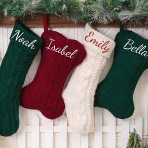 May include: Four cable-knit Christmas stockings hanging in a row. Two are dark green, one is deep red, and one is cream. Each stocking features a personalized name embroidered in white or red script: Noah, Isabel, Emily, and Bella.