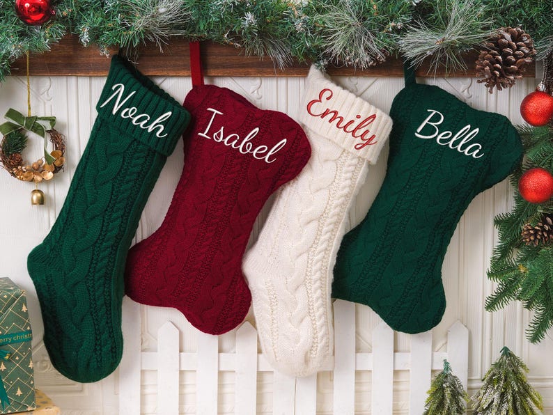 May include: Four cable-knit Christmas stockings hanging in a row. Two are dark green, one is deep red, and one is cream. Each stocking features a personalized name embroidered in white or red script: Noah, Isabel, Emily, and Bella.