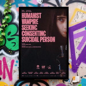 May include: A movie poster with a black background and pink text that reads "Humanist Vampire Seeking Consenting Suicidal Person." The poster features a woman's face and a bottle with a syringe. The poster is framed in black and set against a graffiti wall.