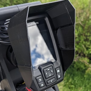 May include: A black sunshade for a Garmin fish finder. The sunshade is made of hard plastic and has a textured surface. The fish finder has a black and white display with a blue sky reflected in it. The fish finder has a series of buttons on the bottom right side.