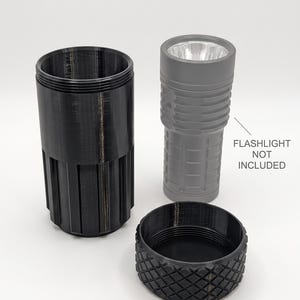 Könnte beinhalten: Ein schwarzer zylindrischer Behälter mit vertikalen Rillen und ein separater Schraubdeckel mit rautenförmiger Textur. Daneben steht eine graue Taschenlampe mit silbernem Reflektor, begleitet vom Text: FLASHLIGHT NOT INCLUDED.
