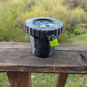 以下が含まれることがあります： 黒い容器で、リブ付きの上面と明るい緑色の南京錠が付いています。「TO TOP 480」という言葉が入ったカラフルなデザインが上面にあります。容器は木製の表面に置かれています。