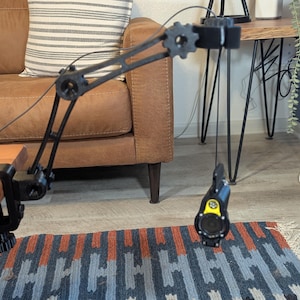 Puede incluir: Un soporte de escritorio negro ajustable con una base de abrazadera, que sostiene un micrófono negro y amarillo. El micrófono está suspendido del soporte por un cable negro.