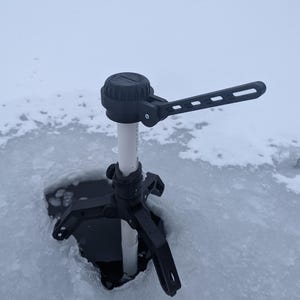 Pode incluir: Um suporte para vara de pesca no gelo preto com um tubo de PVC branco e uma alavanca preta. O suporte está montado em um furo no gelo.