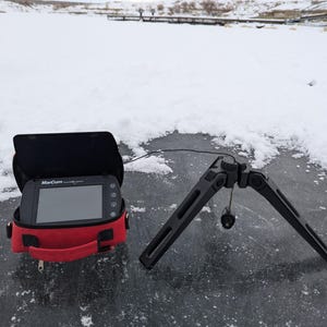 Puede incluir: Un detector de peces MarCum negro en una funda de transporte roja con un trípode negro en un lago helado y nevado.