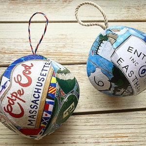 May include: Two handmade Christmas ornaments made from paper. The ornament on the left is blue, white, and red with the text "Cape Cod Massachusetts". The ornament on the right is blue, white, and green with the text "Entering Eastham Inc. 1651".
