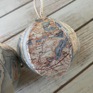 May include: A white, paper mache ornament shaped like a sphere with a map of a lake and surrounding area printed on it. The map is in shades of blue, brown, and red. The ornament has a white twine hanger.