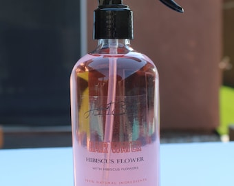 May include: A clear glass bottle with a black spray nozzle filled with pink liquid. The bottle has a label that reads "Hair Water Hibiscus Flower with Hibiscus Flowers 100% Natural Ingredients".