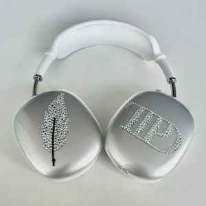 May include: White over-ear headphones with silver earcups. One earcup features a feather design made of rhinestones, the other has the initials "TB" in rhinestones. The headband is white, and the frame is silver.