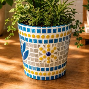 Op de afbeelding: Een cilindervormige plantenbak versierd met een mozaïekpatroon van blauwe, gele en zilveren tegeltjes. De pot heeft een centraal bloemmotief en bevat een levendige paarse orchidee samen met diverse groene planten en bladeren.
