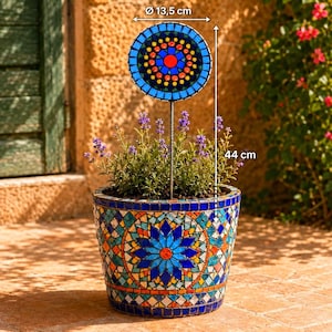 Può includere: Un vaso da fiori in mosaico con un motivo geometrico blu, arancione e bianco contenente una pianta e un picchetto da giardino circolare in mosaico. Il picchetto ha un diametro di 13,5 cm e un'altezza totale di 44 cm, come indicato dalle etichette.