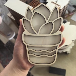 Op de afbeelding: Een lichtgekleurde houten uitsparing in de vorm van een vetplant in een pot. Het ontwerp heeft gelaagde, afgeronde bladeren aan de bovenkant en een potbasis versierd met twee horizontale golvende lijnen. De randen vertonen donkere laser-snijdetails.