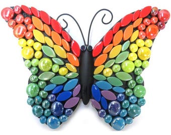 Puede incluir: Un kit de mosaico de mariposa arcoíris. La mariposa está hecha de pequeñas baldosas de vidrio en una variedad de colores, incluyendo rojo, naranja, amarillo, verde, azul, índigo y violeta. La mariposa está montada en un marco de metal negro.