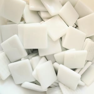 Può includere: Una collezione di numerose piccole tessere quadrate in vetro bianco. Ogni tessera presenta una superficie liscia e lucida con una finitura opaca. Le tessere sono disposte in un mucchio denso e sovrapposto, mostrando la loro forma uniforme.