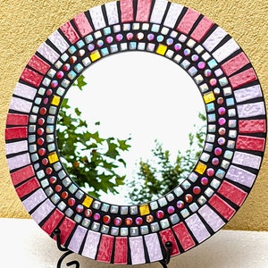 May include: A round mirror with a mosaic frame made of pink, red, and black tiles. The frame is decorated with iridescent glass beads.