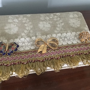 May include: A decorative book cover with a floral pattern in shades of green and white. It features a white lace trim, a gold bow brooch, a butterfly brooch, a pearl brooch, and gold fringe.