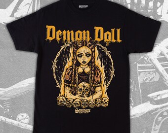 May include: Black t-shirt with a graphic print of a girl with a skull in her hands. The girl is surrounded by flames and barbed wire. The text "Demon Doll" is printed above the graphic. The text "Scourge Cartel" is printed below the graphic.