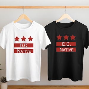 May include: Two t-shirts, one white and one black, each with a red design. The design features three red stars above a red horizontal bar with the text "D.C. NATIVE" in white letters. Both shirts are hanging on wooden hangers.