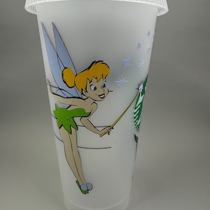 May include: A clear plastic Starbucks cold cup with a lid, featuring a colorful illustration of Tinkerbell. The design includes Tinkerbell with her wand, blue wings, and a green dress. The Starbucks logo is also visible.