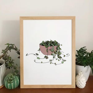 May include: A framed art print depicting a pink plant pot filled with green foliage and trailing vines. The artwork is set against a white background and framed in light wood. The plant has small, heart-shaped leaves.