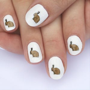 May include: Manicured nails with a white base featuring brown rabbit designs. The rabbits are depicted in a cartoon style, with a light brown body and a darker brown ear. The nails are oval-shaped.
