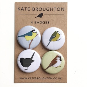 May include: Four round badges featuring bird illustrations. The badges are on a brown card with the text "KATE BROUGHTON" and "4 BADGES". The website address is also printed on the card. The bird illustrations include a blue tit, a great tit, a black bird, and a sparrow.