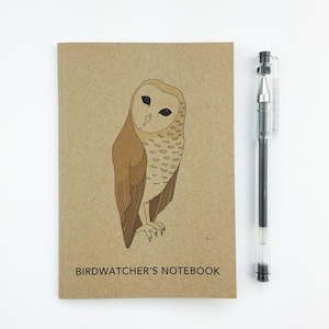 May include: A brown Birdwatcher's Notebook with an illustration of a barn owl. The notebook is next to a clear pen with black ink. The text "Birdwatcher's Notebook" is printed at the bottom of the notebook.
