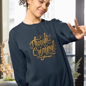 Peut inclure: Sweat-shirt bleu marine avec l'inscription "Thought Criminal" en lettres dorées et ornées. Le sweat-shirt a un col rond et des manches longues. Le design est centré sur la poitrine.
