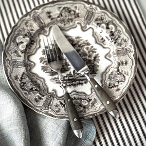May include: A decorative plate with a black and white landscape pattern, alongside a fork and a knife. The cutlery features dark wooden handles and silver details. The plate and utensils are placed on a striped fabric and a folded grey cloth.